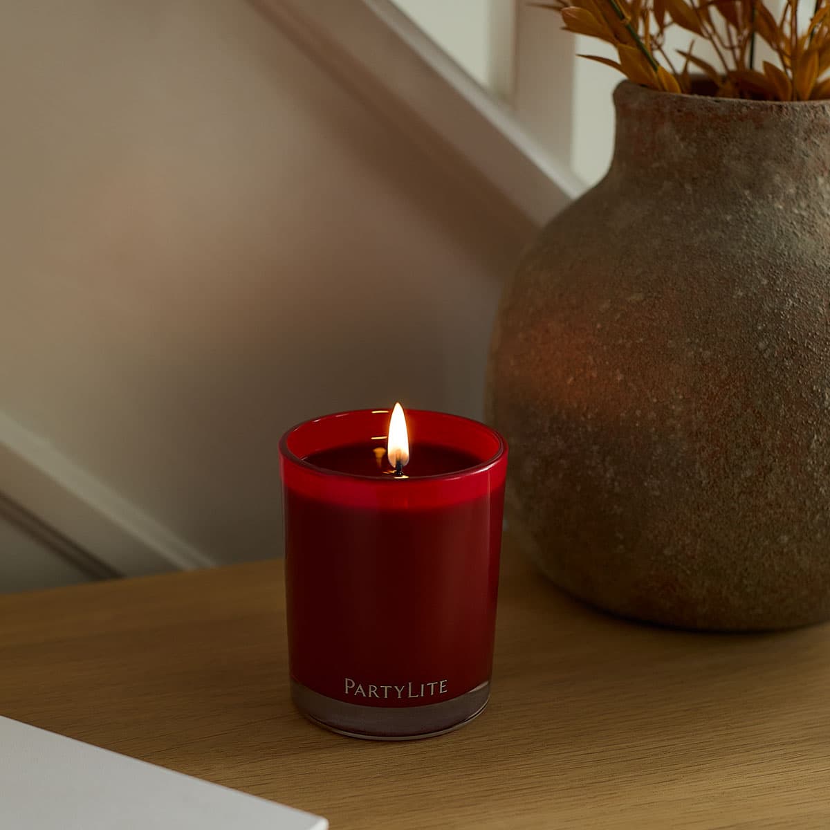 Autumn Sangria Escential Jar lit on a wooden surface with a vase in the background.