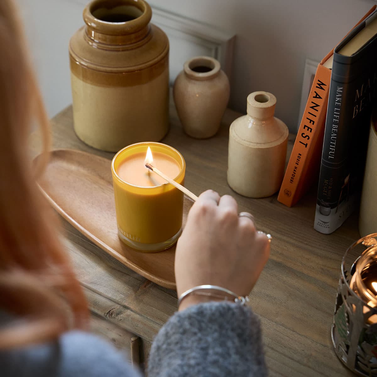 Whiskey & Oak Escential Jar being lit by a lady with a long matchstick on a wooden surface with ceramic vases and books in the background.