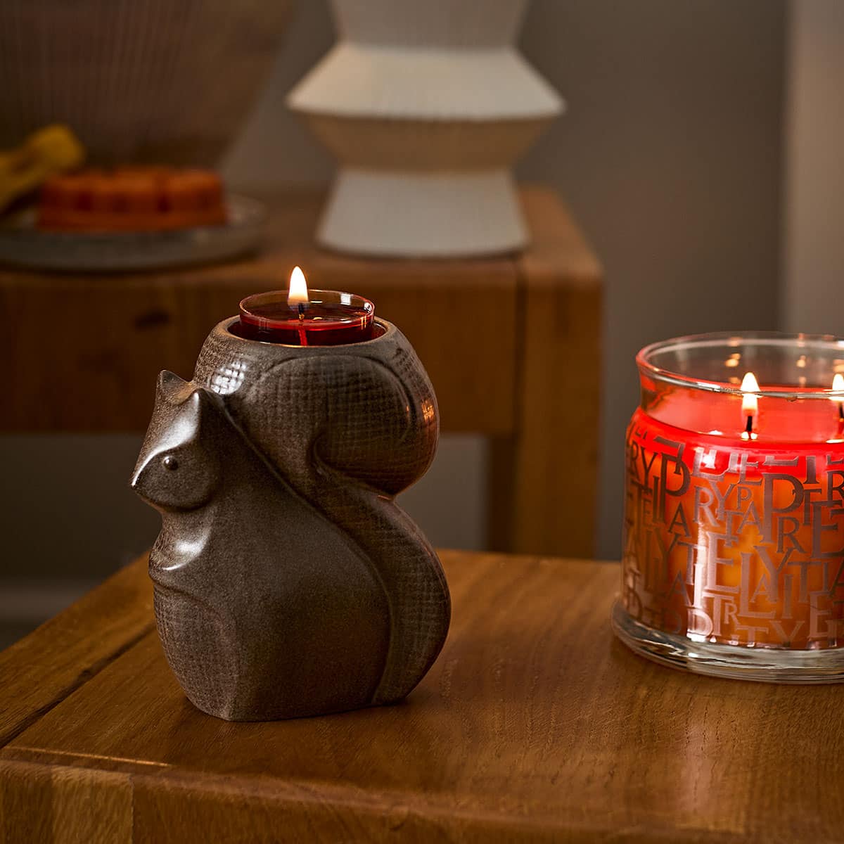 Forest Friends Squirrel Tealight Holder shown with a lit tealight beside a lit 3-wick jar candle on a wooden surface.