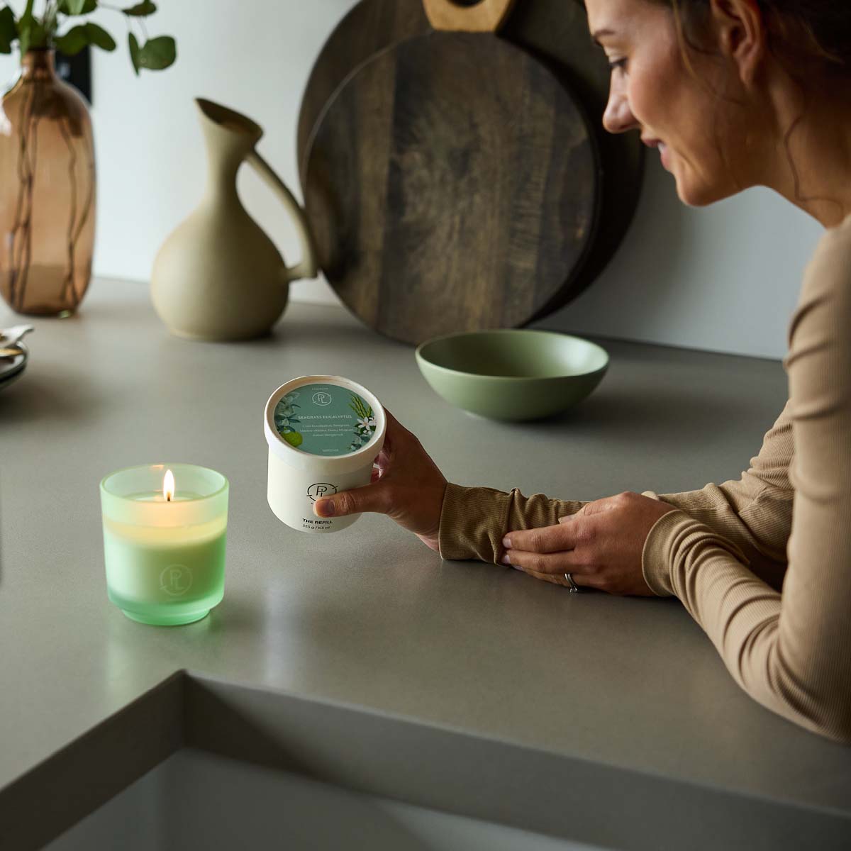 Seagrass Eucalyptus Wax Refill shown with the Seagrass Eucalyptus Refillable jar lit on a counter with a lady holding the refill.