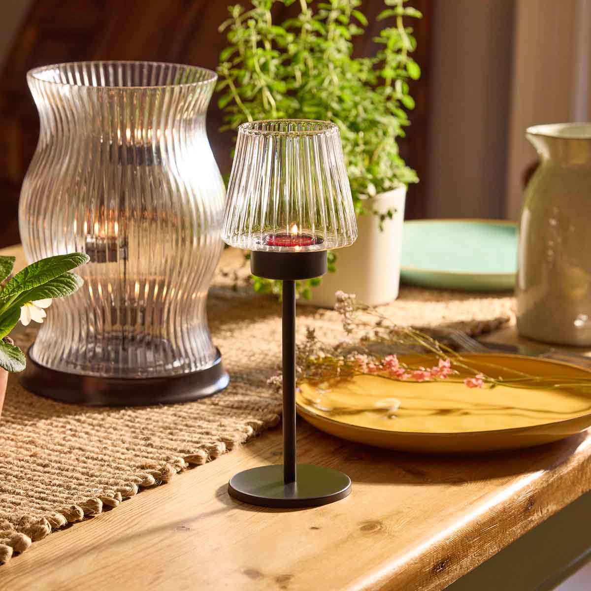Modern Farmhouse Tealight Lamp shown on a wooden table top with hurricane and house plant in the background.