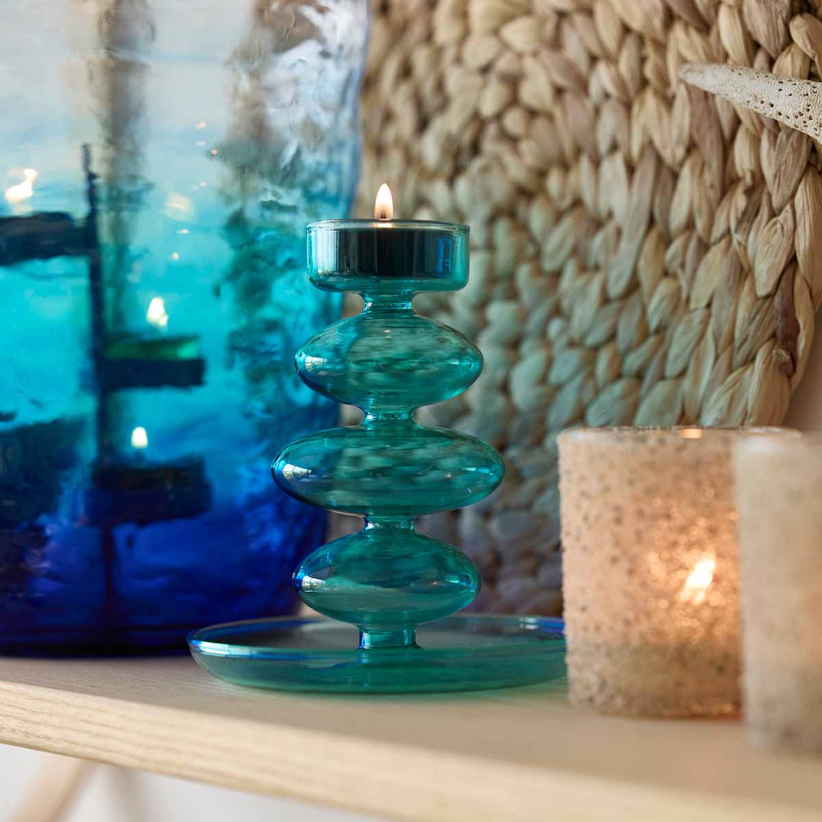 Blue Waves Tealight Holder with a lit tealight against a blue Hurricane with a woven mat in the background.