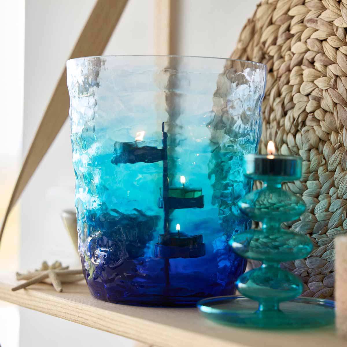 Ombre Waves Hurricane with 3 lit candles on a metal tealight tree with shades of blue and green with a woven table mat in the background.