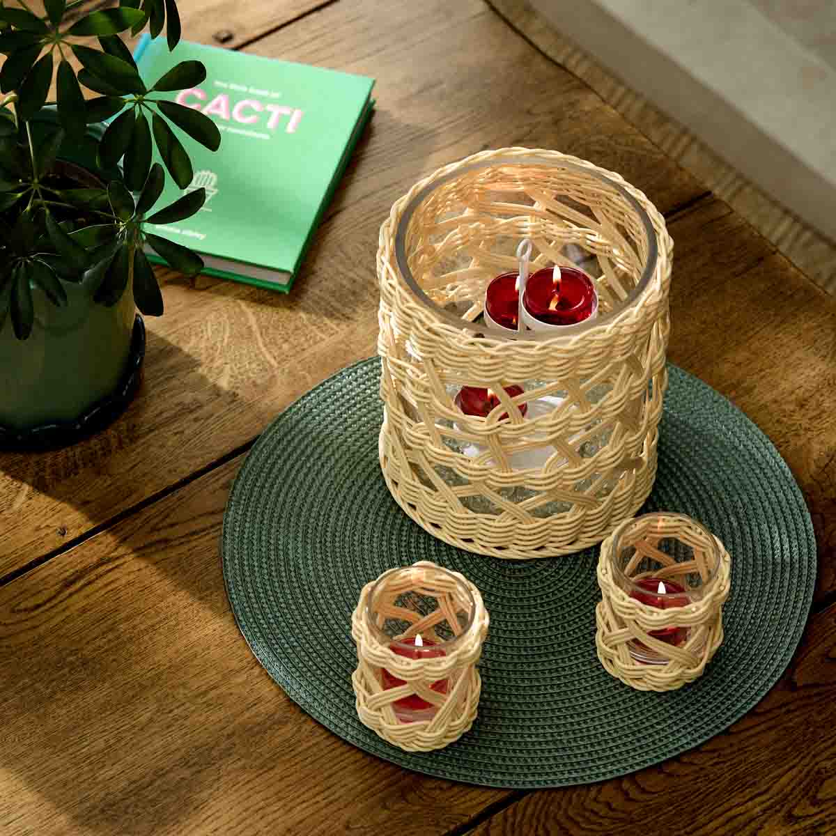 Woven Tealight/Votive Holder and the Woven Hurricane on a green table mat on a wooden table with a plant and book in the background.