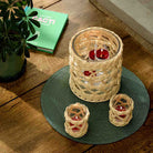 Woven Tealight/Votive Holder and the Woven Hurricane on a green table mat on a wooden table with a plant and book in the background.