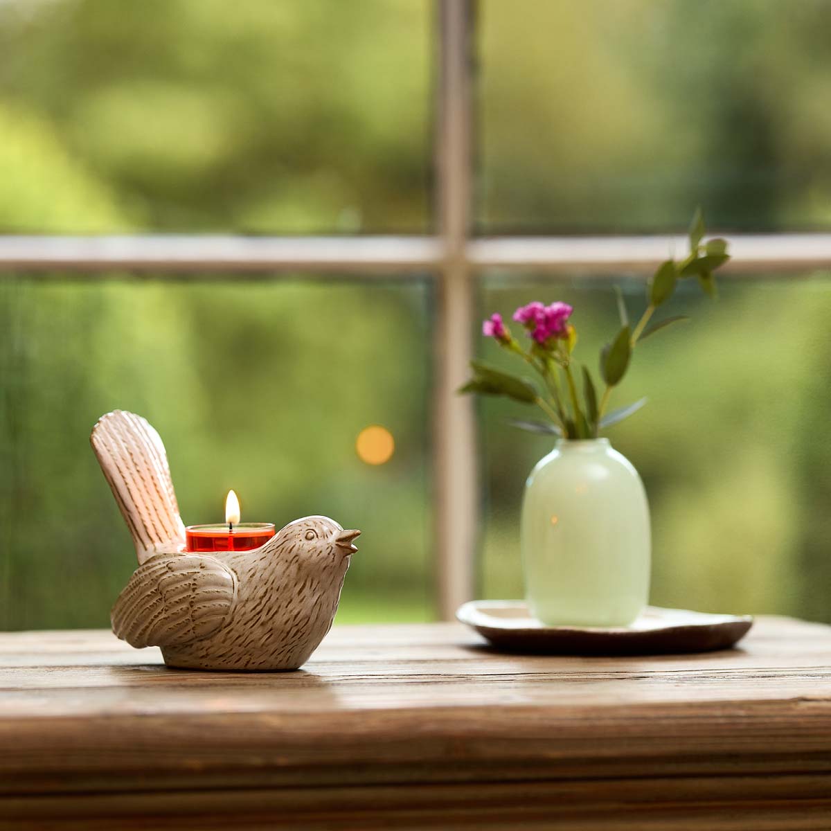 Songbird Tealight Holder shown on a wooden surface with a vase of flowers and a window in the background.
