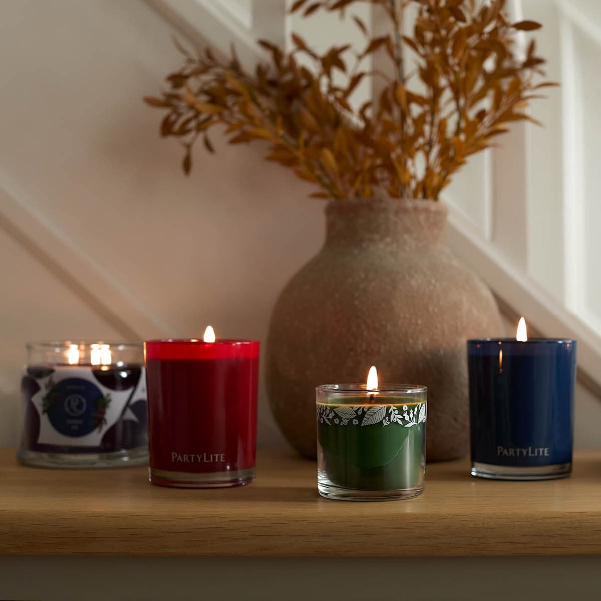 Escential Jar Candles, Mini Jar Candle and a 3-wick Jar Candle all lit against a wooden surface with a vase of leaves in the background.