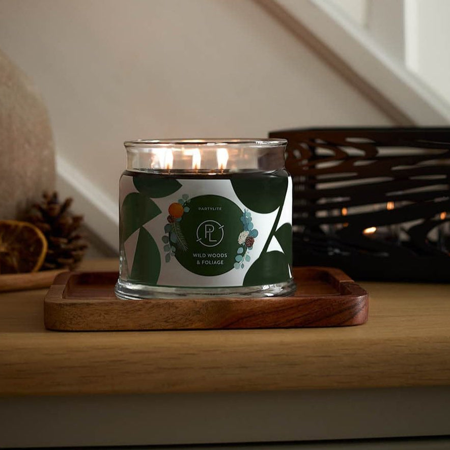 Wild Woods & Foliage 3-Wick Jar Candle lit on a wooden surface with white stair bannisters in the background.