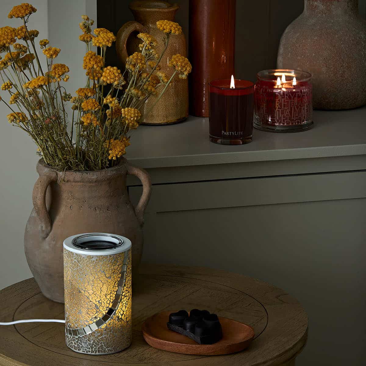 Modern Crushed Mosaic Electric Wax Melt Warmer shown on a wooden round table with a pottery vase with flowers and lit candles in the background.
