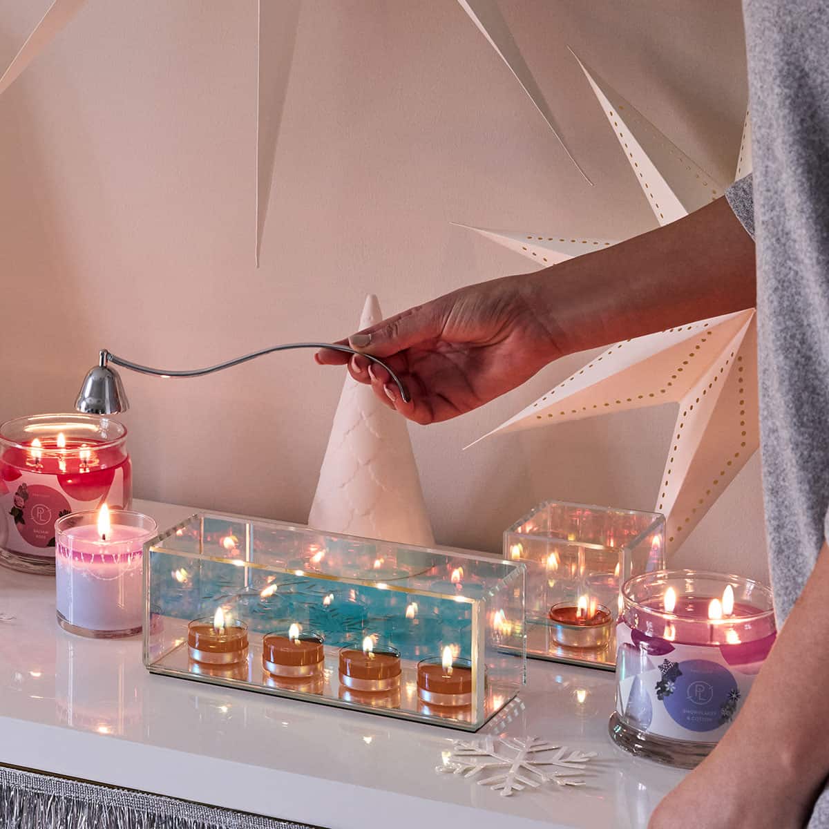 Infinite Reflections Centerpiece shown with 4 lit candles, a Mini Jar candle and 3-wick jar candles also lit with a lady's hand holding a chrome snuffer.