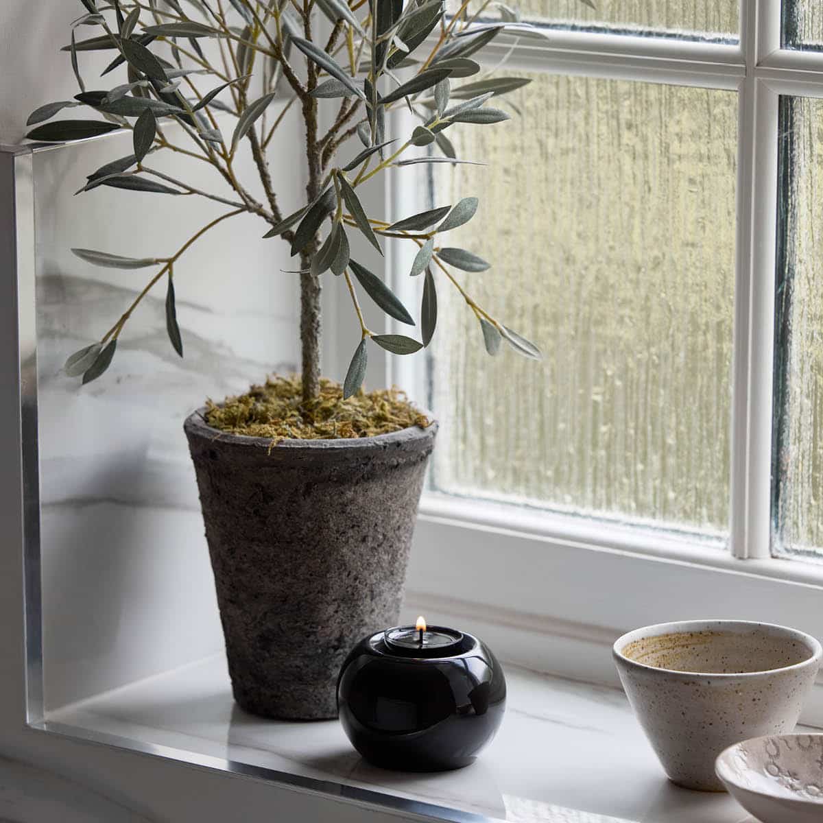 Tranquil Orb Tealight Holder with a lit tealight displayed on a light colored windowsill next to a house plant.