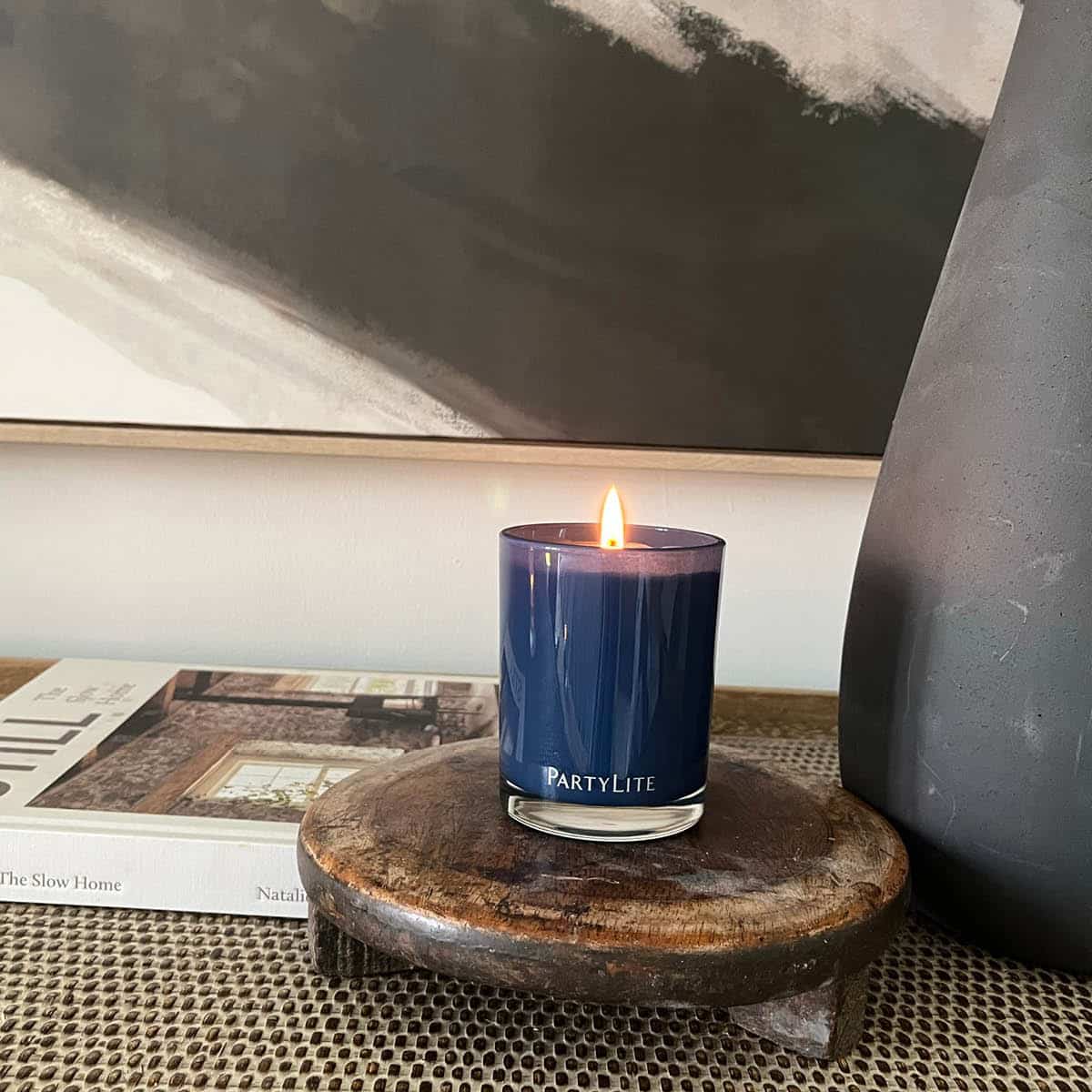 Nordic Air Escential Jar candle lit on a wooden coaster on a table top, with a book in the background.