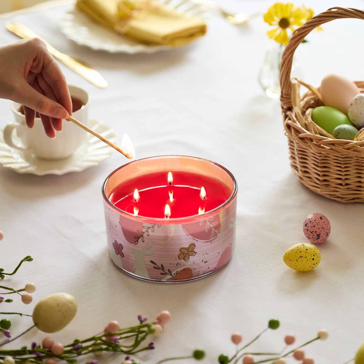 Garden Party 4-Wick Jar Candle being lit by a long match, beside a basket of Easter Eggs on a white table cloth.