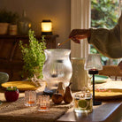 Modern Farmhouse Hurricane shown with the Modern Tealight Lamp and other tealight holders with a lit 3-wick on a table with a woven table runner with house plant in the background.
