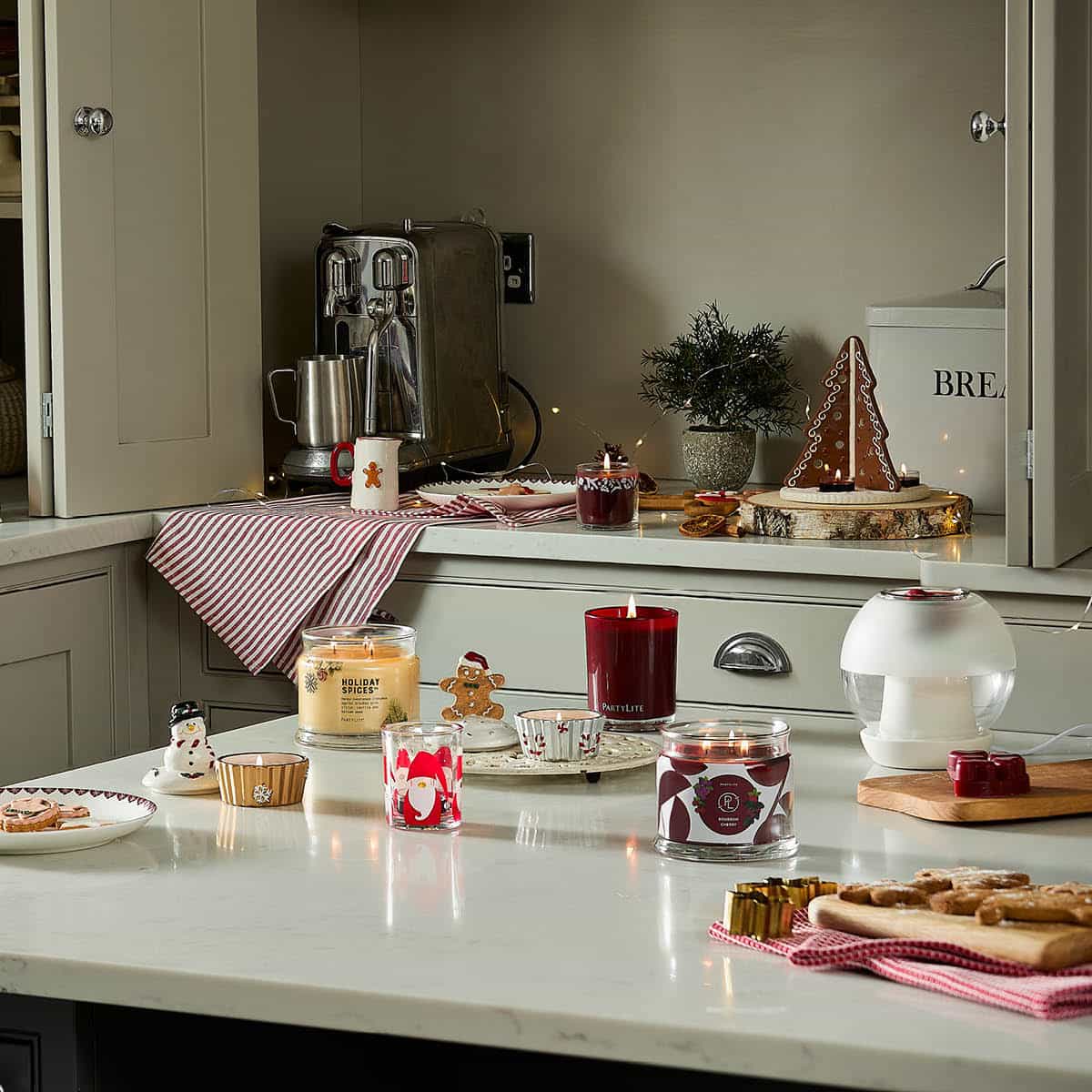 An assortment of PartyLite items ranging from Escential jar, 3-wick candles, festive tealight holders on a kitchen counter with kitchen items in the background.