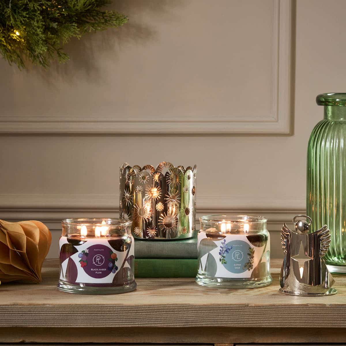 two 3-wick jar candles lit with a Angel Tealight holder and a silver holder in the background on a wooden surface.