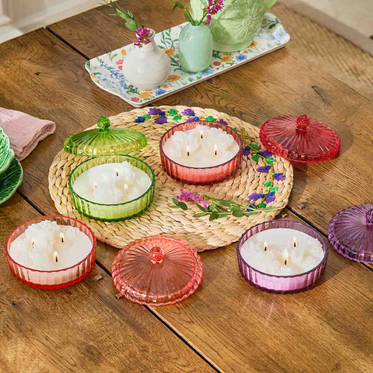 Assortment of Garden Party 3-Wick colorful Jar Candles on a woven table mat on a wooden table with decorative items in the background.