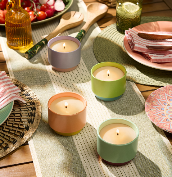 Four Citronella single wick jar candles in duo color holders on a woven table mat surrounded by plates and cutlery on a wooden table