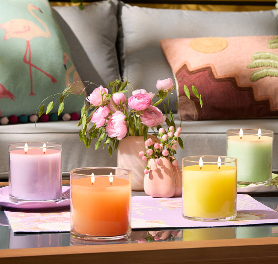Assorted Colorful 2 wick GloLite candle jars on a glass table with vases of flowers and a sofa in the background