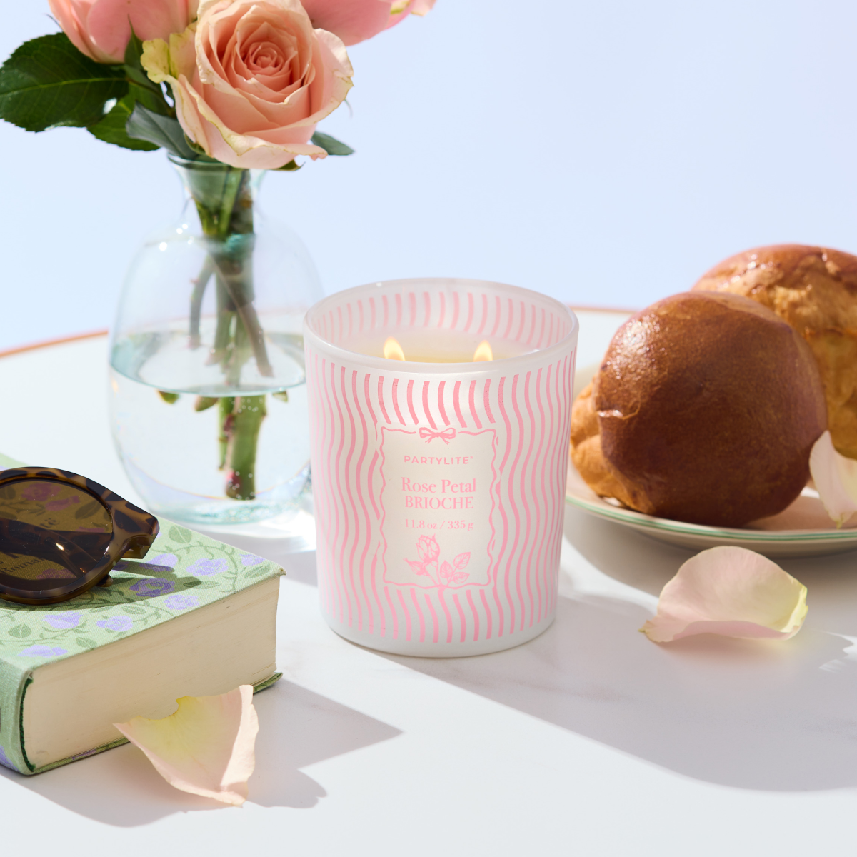 Rose Petal Brioche jar candle, with pink and white waves on a white table with vase and flowers and a book with sunglasses.