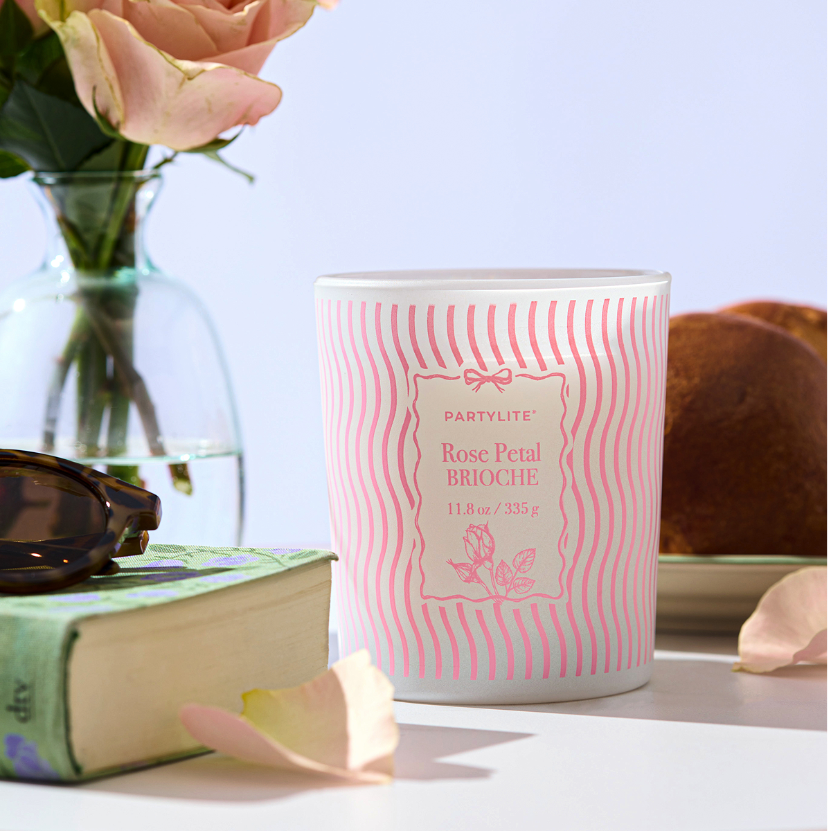 Rose Petal Brioche jar candle on a surface with a book, sunglasses, and flowers in the background.
