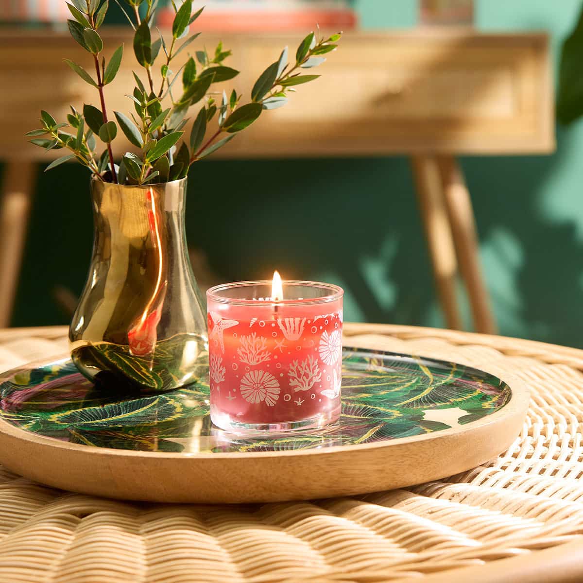 Beachside Tiare Mini Jar Candle on a leaf design wooden tray next to a metallic vase of leaves on a woven table top with a green background
