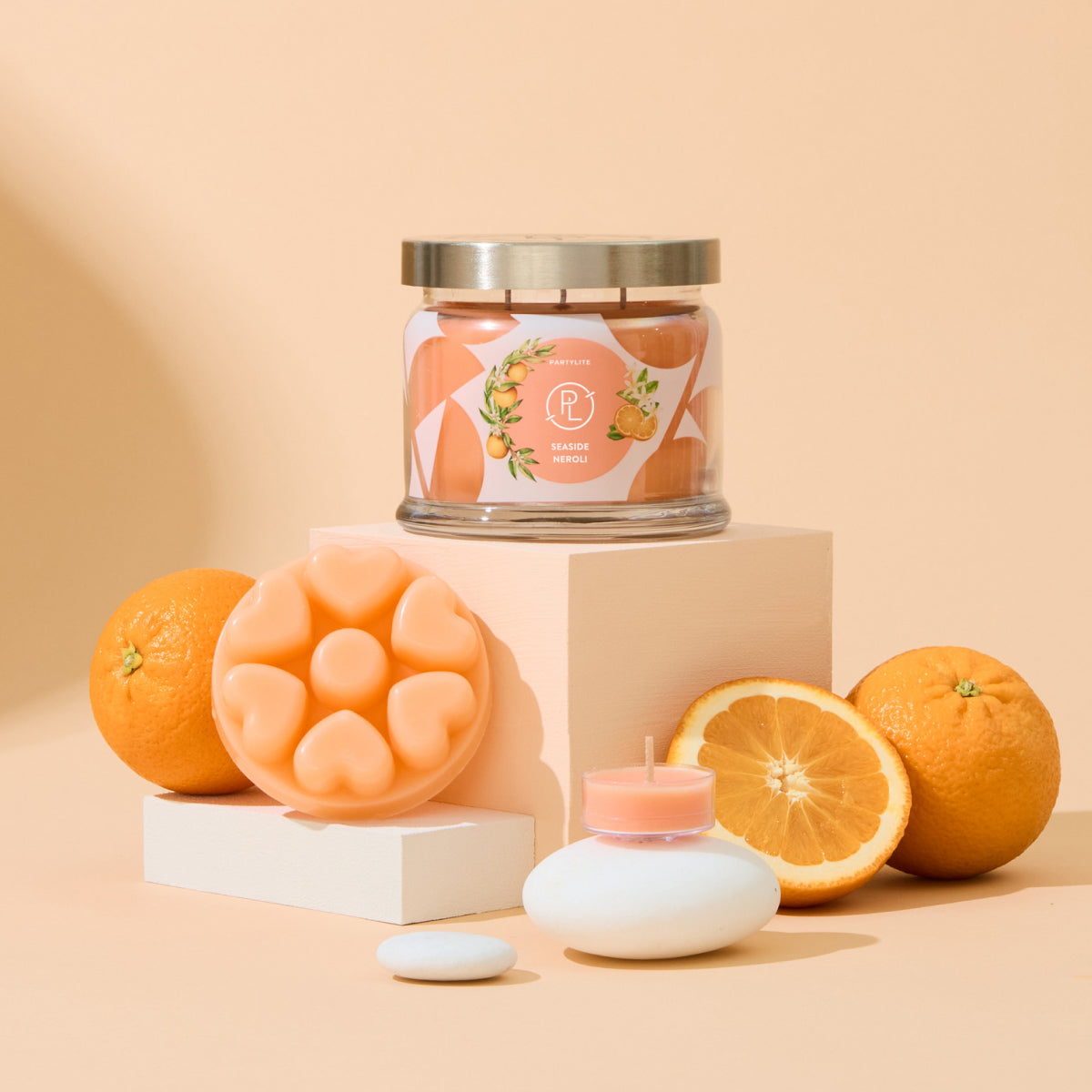 Seaside Neroli fragrance, shown in 3-wick jar candle, heart shaped wax melts and a tealight, with oranges against a light orange background.