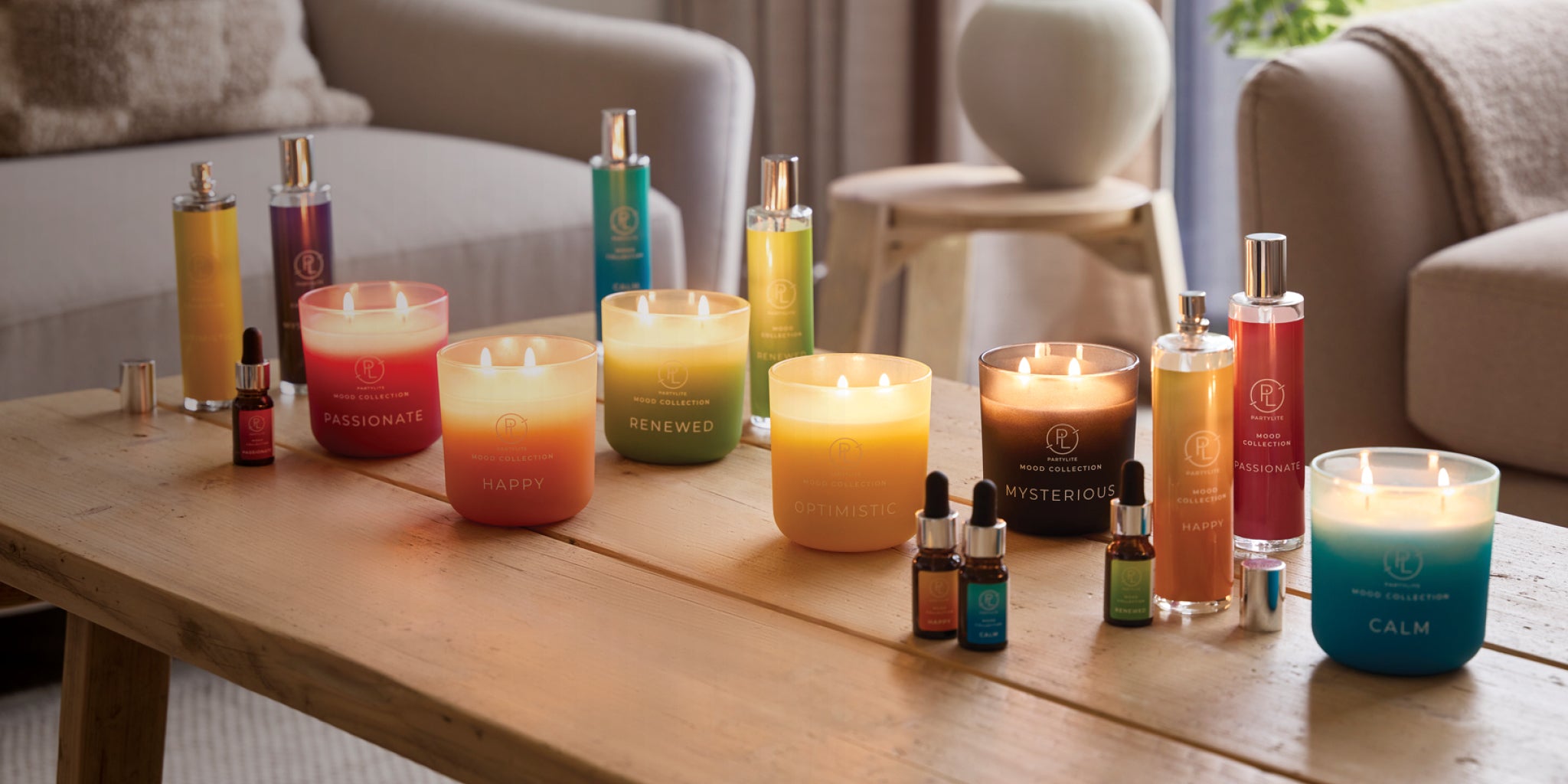 Assorted Mood Collection items. Jar Candles, Room Sprays, Fragranced Oils, and Smart Home Plug-in Refills. All displayed on a wooden table with a living room background