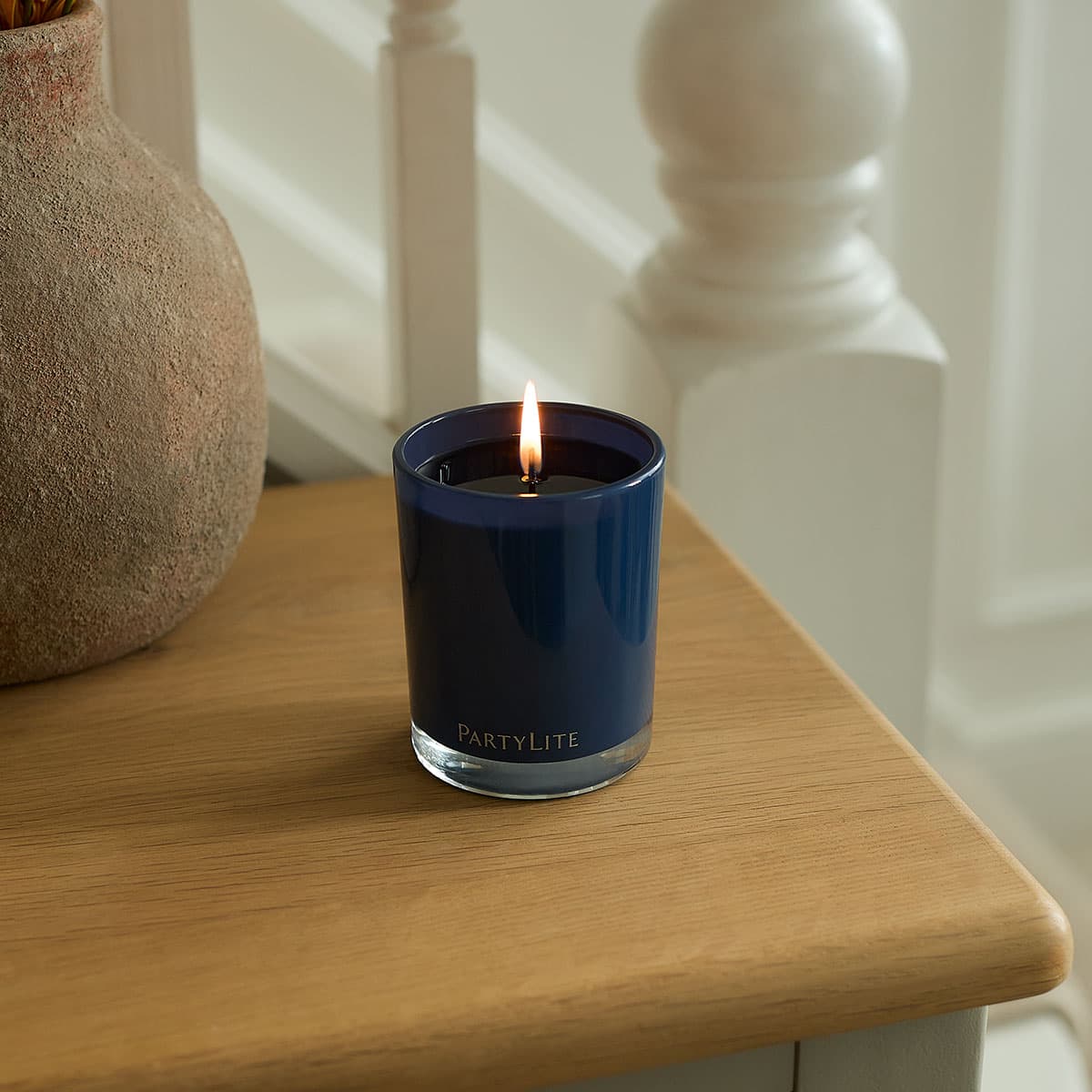 Nordic Air Escential Jar Candle in a blue color lit on a wooden surface, with white stair bannisters in the background.
