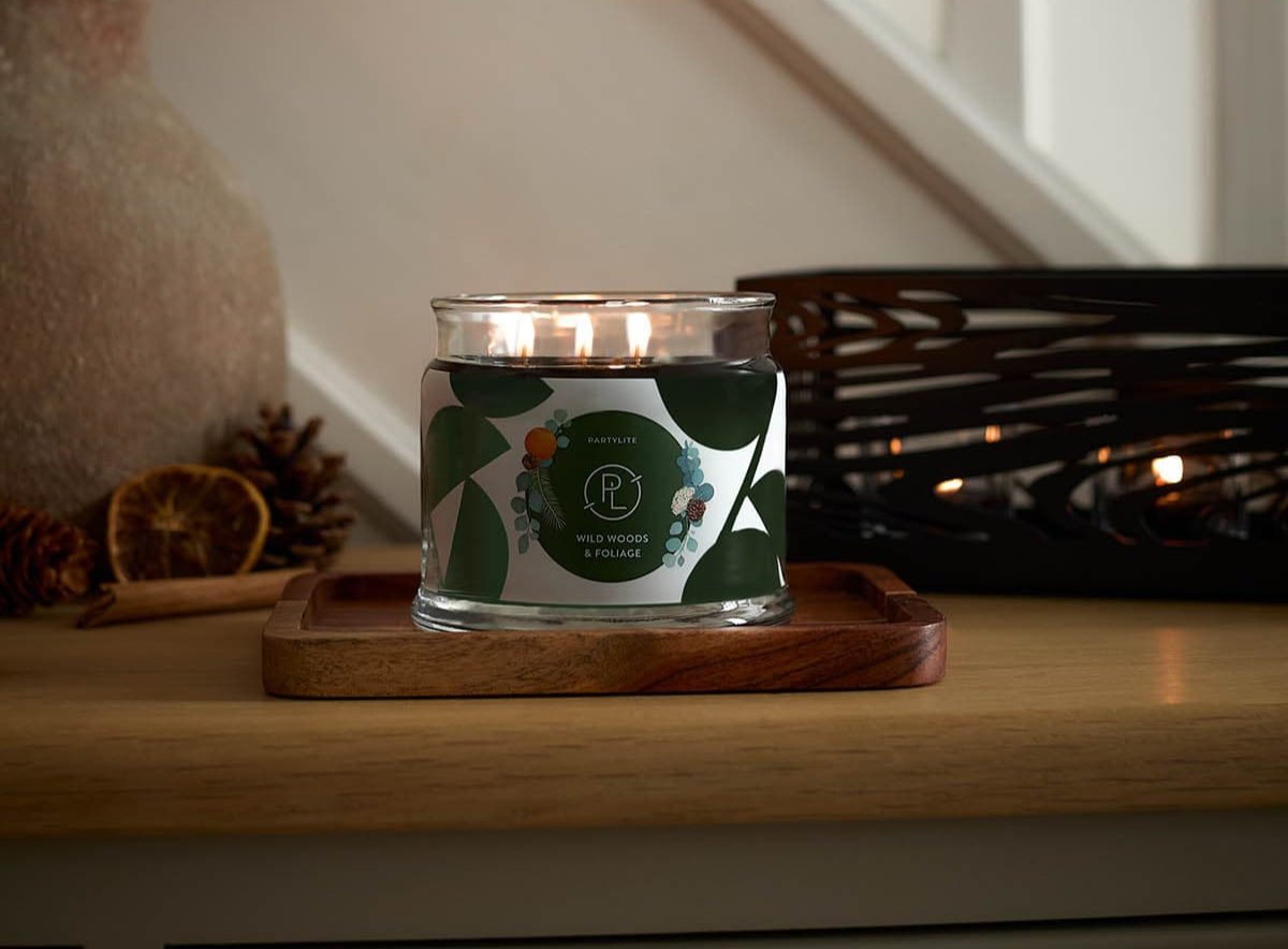 Wild Woods & Foliage 3-Wick Jar Candle lit on a wooden surface with white stair bannisters in the background.