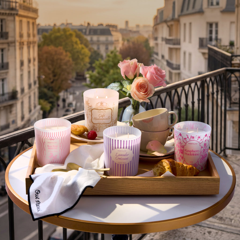 Wooden tray displaying four Cafè Patisserie candles with dishes and a vase with flowers on a Parisian cafe table on balcony with background of the city