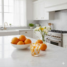 Happy Fragrance Marbled jar candle on a kitchen worktop with a bowl of oranges and a clear vase of white flowers next to it.
