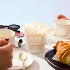 Candle labeled 'Cafe Madeleine' on a table with cups, croissant, and sunglasses.